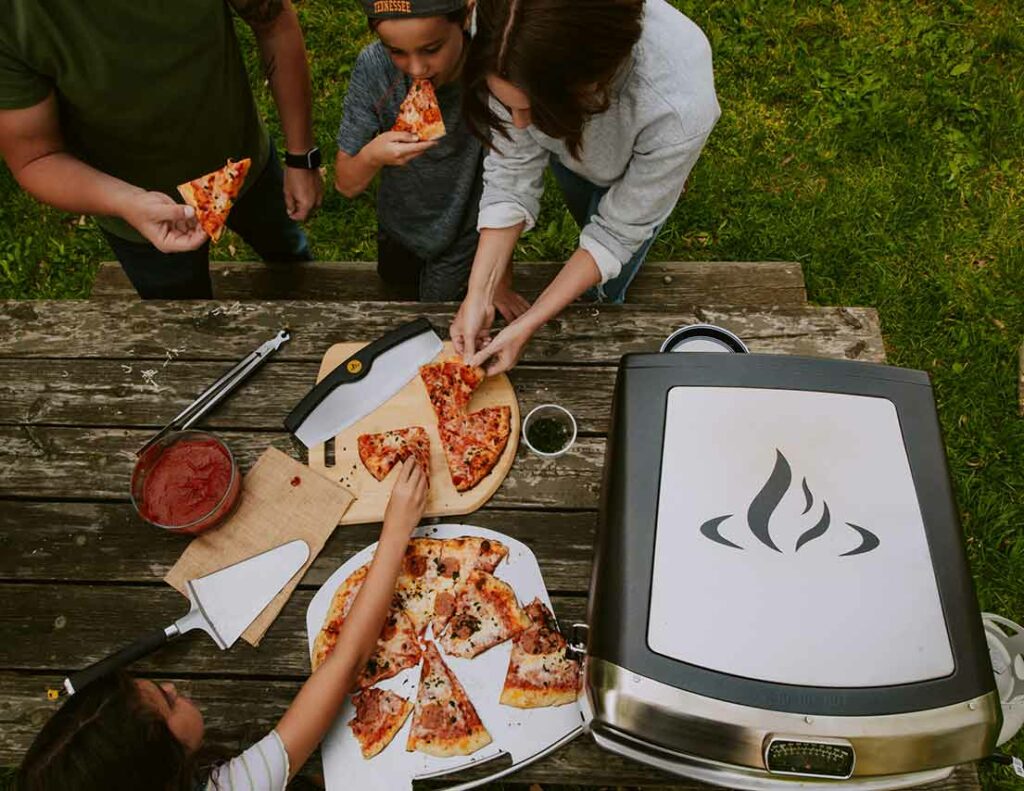 Family at a picnic enjoying freshly cooked pizza from a HALO Versa Pizza Oven. Daytime. Dining al fresco.